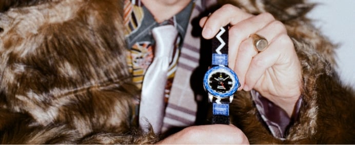 A man in a fur coat holds the Limited Edition Zodiac x Rowing Blazers Super Sea Wolf GMT watch featuring a blue bezel denoting 27 global cities.